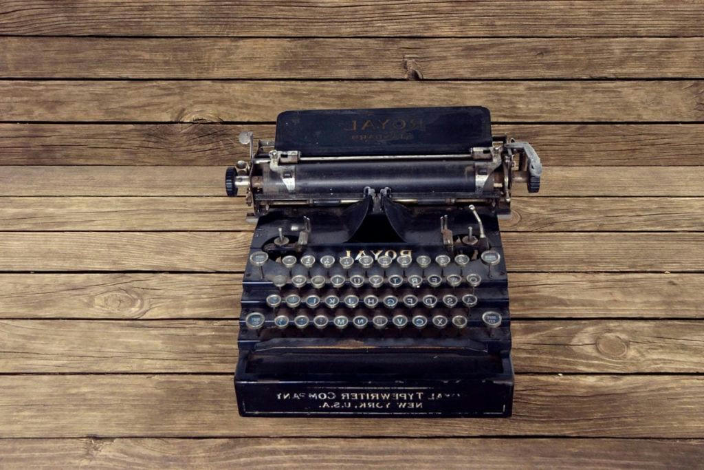 This image shows an old, vintage typewriter placed on a wooden surface. The typewriter is black and has round keys with characters printed on them. The brand “ROYAL” is visible at the top of the typewriter. It’s placed on a surface made of horizontal wooden planks, showcasing a rustic aesthetic. There are visible signs of wear and age on both the typewriter and the wooden surface, giving it an antique look.